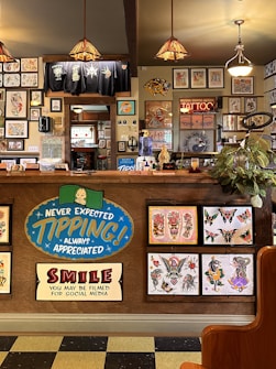 The image depicts the interior of a tattoo parlor. Artwork of various tattoo designs is displayed on the walls and a wooden counter. The counter features colorful signs related to tipping and social media. There are pendant lights with stained glass shades hanging from the ceiling, and a variety of framed pictures and designs provide a vibrant backdrop. A water cooler is present on the counter, and a collection of plants adds a touch of greenery.