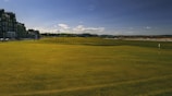 A wide expanse of a golf course with neatly trimmed green grass under a clear blue sky. On the left side, a row of historic buildings lines the perimeter, adding a classic architectural touch. The horizon is dotted with distant hills and a few scattered clouds.