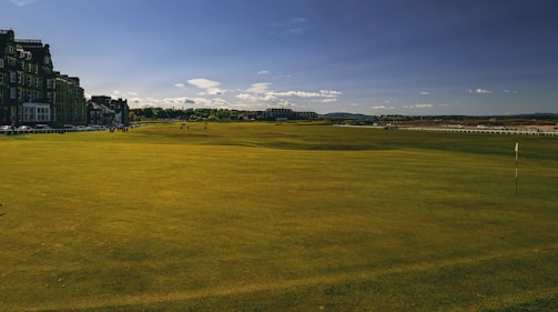A wide expanse of a golf course with neatly trimmed green grass under a clear blue sky. On the left side, a row of historic buildings lines the perimeter, adding a classic architectural touch. The horizon is dotted with distant hills and a few scattered clouds.