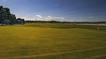 A wide expanse of a golf course with neatly trimmed green grass under a clear blue sky. On the left side, a row of historic buildings lines the perimeter, adding a classic architectural touch. The horizon is dotted with distant hills and a few scattered clouds.