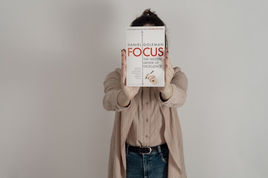 A person holding a book titled 'Focus: The Hidden Driver of Excellence' by Daniel Goleman in front of their face. The background is a plain light color, and the person is wearing a beige shirt and jeans.