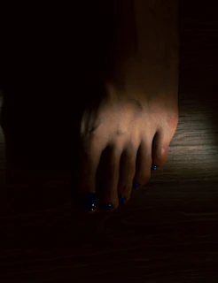 A human foot with toenails painted in a dark blue color is visible. The foot is set against a dark background with minimal lighting, creating shadows that obscure parts of the image.