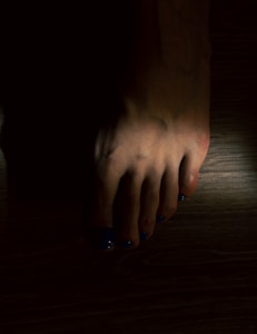 A human foot with toenails painted in a dark blue color is visible. The foot is set against a dark background with minimal lighting, creating shadows that obscure parts of the image.