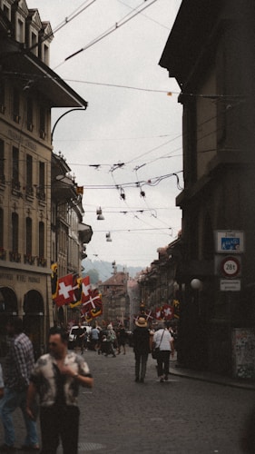 A street scene with people walking along a cobblestone road flanked by tall buildings with arched windows. Swiss flags and banners are visible, and the street is adorned with hanging overhead wires. The atmosphere appears to be lively with people dressed casually walking towards the horizon where the landscape softens into a distant haze.