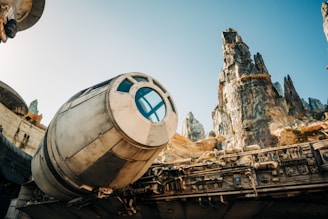 A futuristic spaceship with a cylindrical cockpit and intricate mechanical details is positioned beneath rugged, towering rock formations against a clear blue sky.