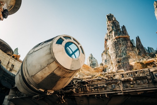 A futuristic spaceship with a cylindrical cockpit and intricate mechanical details is positioned beneath rugged, towering rock formations against a clear blue sky.