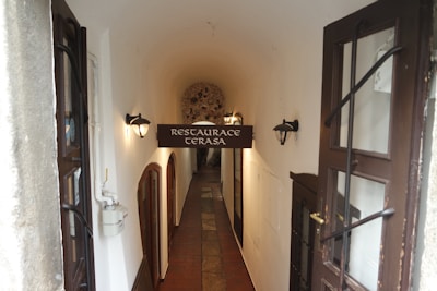 An arched hallway leads to a cozy restaurant entrance with a sign reading 'Restaurace Terasa'. The corridor features dim wall-mounted lights, wooden doors, and a tiled floor, creating a warm and inviting ambiance.