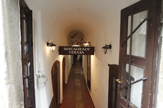 An arched hallway leads to a cozy restaurant entrance with a sign reading 'Restaurace Terasa'. The corridor features dim wall-mounted lights, wooden doors, and a tiled floor, creating a warm and inviting ambiance.