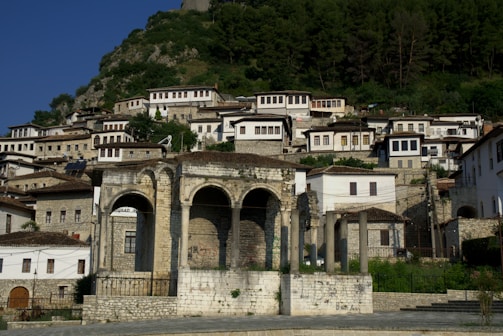 A hillside town with traditional Ottoman-style stone houses and a large ancient stone structure with arches in the foreground. The houses have red-tiled roofs and wooden window frames, nestled into the green hillside.