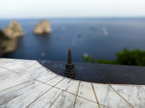 A sundial made of white marble and metal is placed on a ledge overlooking a scenic ocean view. In the background, two large rocky formations are visible, partially blurred to bring focus to the sundial in the foreground. The ocean is calm with a few scattered boats.