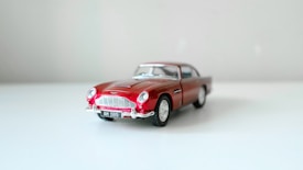 A detailed model of a classic red car is placed on a clean, light surface. The car features intricate details, including realistic wheels, a shiny finish, and a distinct front grille. The background is plain and neutral, enhancing the focus on the car model.