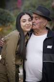 A young woman with long dark hair is affectionately leaning into an elderly man. The man is wearing a black hat, black Nike jacket, and white t-shirt. They appear to be outdoors with blurred greenery in the background.