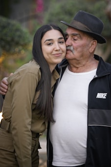 A young woman with long dark hair is affectionately leaning into an elderly man. The man is wearing a black hat, black Nike jacket, and white t-shirt. They appear to be outdoors with blurred greenery in the background.