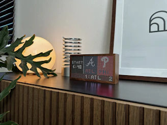 A modern interior scene featuring a wooden cabinet with a dark countertop. On the surface, there is a digital clock with a pixelated display in a wooden frame, a decorative coil sculpture, a round luminous lamp casting a warm glow, and a leafy plant partly covering the lamp. A framed abstract artwork leans against the wall.