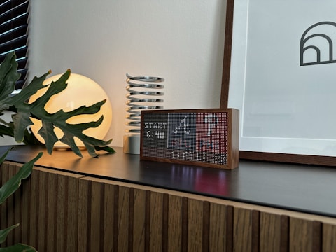 A modern interior scene featuring a wooden cabinet with a dark countertop. On the surface, there is a digital clock with a pixelated display in a wooden frame, a decorative coil sculpture, a round luminous lamp casting a warm glow, and a leafy plant partly covering the lamp. A framed abstract artwork leans against the wall.