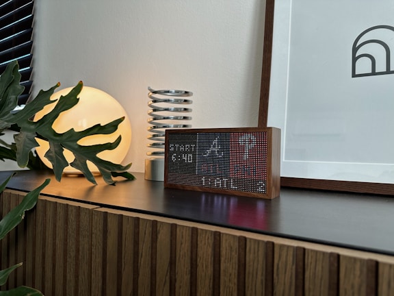 A modern interior scene featuring a wooden cabinet with a dark countertop. On the surface, there is a digital clock with a pixelated display in a wooden frame, a decorative coil sculpture, a round luminous lamp casting a warm glow, and a leafy plant partly covering the lamp. A framed abstract artwork leans against the wall.