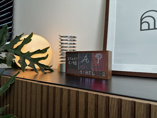 A modern interior scene featuring a wooden cabinet with a dark countertop. On the surface, there is a digital clock with a pixelated display in a wooden frame, a decorative coil sculpture, a round luminous lamp casting a warm glow, and a leafy plant partly covering the lamp. A framed abstract artwork leans against the wall.