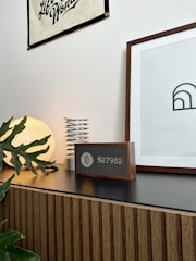 A modern interior setting featuring a wooden cabinet with horizontal grooves. On top of the cabinet, there is a round lamp emitting a warm glow, a metallic spring-like object, and a digital clock or speaker with a wooden frame. A framed minimalist art piece and a wall banner are mounted on the white wall, while a few green plant leaves add a touch of nature to the scene.