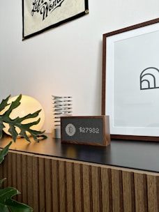 A modern interior setting featuring a wooden cabinet with horizontal grooves. On top of the cabinet, there is a round lamp emitting a warm glow, a metallic spring-like object, and a digital clock or speaker with a wooden frame. A framed minimalist art piece and a wall banner are mounted on the white wall, while a few green plant leaves add a touch of nature to the scene.