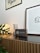 A modern interior setting featuring a wooden cabinet with horizontal grooves. On top of the cabinet, there is a round lamp emitting a warm glow, a metallic spring-like object, and a digital clock or speaker with a wooden frame. A framed minimalist art piece and a wall banner are mounted on the white wall, while a few green plant leaves add a touch of nature to the scene.