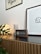 A modern interior setting featuring a wooden cabinet with horizontal grooves. On top of the cabinet, there is a round lamp emitting a warm glow, a metallic spring-like object, and a digital clock or speaker with a wooden frame. A framed minimalist art piece and a wall banner are mounted on the white wall, while a few green plant leaves add a touch of nature to the scene.