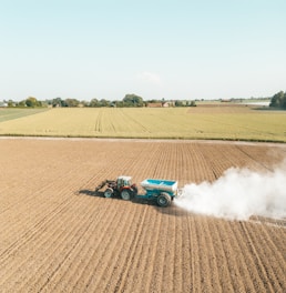 A tractor is spreading fertilizer or lime on a vast, freshly plowed agricultural field. The fields stretch towards the horizon, bordered by trees and distant farm buildings under a clear blue sky. White dust billows behind the tractor, contrasting with the soil's earthy tones.