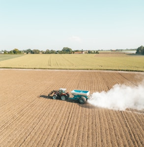 A tractor is spreading fertilizer or lime on a vast, freshly plowed agricultural field. The fields stretch towards the horizon, bordered by trees and distant farm buildings under a clear blue sky. White dust billows behind the tractor, contrasting with the soil's earthy tones.
