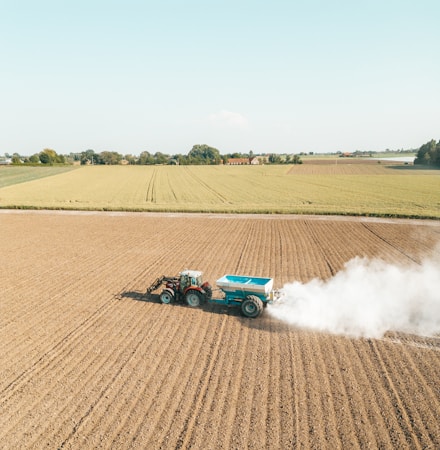 A tractor is spreading fertilizer or lime on a vast, freshly plowed agricultural field. The fields stretch towards the horizon, bordered by trees and distant farm buildings under a clear blue sky. White dust billows behind the tractor, contrasting with the soil's earthy tones.
