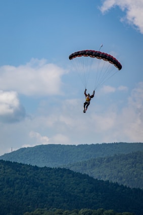 A person is paragliding through a clear blue sky, with lush green mountains in the background. The parachute is prominently visible with its dark colors contrasting against the sky.