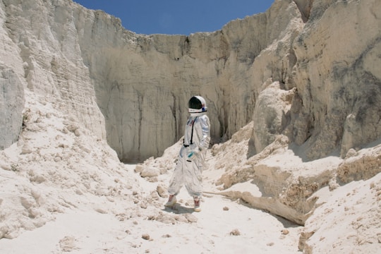 An astronaut in a white spacesuit stands amid rocky, pale sand cliffs, suggesting a moon-like landscape under a clear blue sky. The terrain is rugged and barren, with various rock formations and a sense of isolation and exploration.
