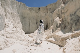 An astronaut in a white spacesuit stands amid rocky, pale sand cliffs, suggesting a moon-like landscape under a clear blue sky. The terrain is rugged and barren, with various rock formations and a sense of isolation and exploration.