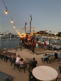 A festive scene at a beachside setting featuring a pirate-themed ship bar named 'Happy Arrr'. It is docked near a sandy area with string lights hanging above, illuminating several people sitting and standing nearby. A variety of boats are visible in the calm water in the background as evening sets in.