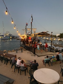 A festive scene at a beachside setting featuring a pirate-themed ship bar named 'Happy Arrr'. It is docked near a sandy area with string lights hanging above, illuminating several people sitting and standing nearby. A variety of boats are visible in the calm water in the background as evening sets in.
