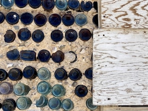 An artistic arrangement of various blue and green glass bottles embedded upside-down in a concrete surface, alongside a wooden textured panel. The bottles create a pattern with their circular bottoms mostly visible, casting shadows and adding depth.