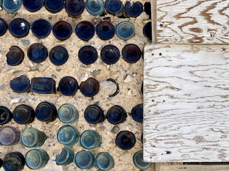 An artistic arrangement of various blue and green glass bottles embedded upside-down in a concrete surface, alongside a wooden textured panel. The bottles create a pattern with their circular bottoms mostly visible, casting shadows and adding depth.