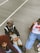 Three people are sitting on a concrete surface outdoors, dressed in casual, trendy streetwear. One person wears a leopard print bucket hat with sunglasses, a black and white varsity jacket, and beige pants. Another person has a red afro hairstyle, a burgundy patterned sweater, and light blue jeans. The third person is wearing a white basketball jersey with red accents and blue jeans.