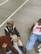Three people are sitting on a concrete surface outdoors, dressed in casual, trendy streetwear. One person wears a leopard print bucket hat with sunglasses, a black and white varsity jacket, and beige pants. Another person has a red afro hairstyle, a burgundy patterned sweater, and light blue jeans. The third person is wearing a white basketball jersey with red accents and blue jeans.