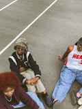 Three people are sitting on a concrete surface outdoors, dressed in casual, trendy streetwear. One person wears a leopard print bucket hat with sunglasses, a black and white varsity jacket, and beige pants. Another person has a red afro hairstyle, a burgundy patterned sweater, and light blue jeans. The third person is wearing a white basketball jersey with red accents and blue jeans.