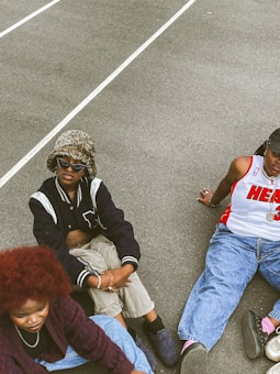 Three people are sitting on a concrete surface outdoors, dressed in casual, trendy streetwear. One person wears a leopard print bucket hat with sunglasses, a black and white varsity jacket, and beige pants. Another person has a red afro hairstyle, a burgundy patterned sweater, and light blue jeans. The third person is wearing a white basketball jersey with red accents and blue jeans.