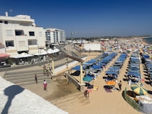 A sandy beach filled with numerous rowed beach umbrellas and sunbeds stretching along the coastline. Several people are visible enjoying the beach. On the left side, modern apartment buildings with outdoor cafes and white umbrellas line the promenade. The sky is clear with no clouds.