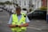 A middle-aged person wearing a high-visibility vest is focused on a device, possibly a phone or a communication device, while standing on a street. The person appears to be involved in official duties, suggested by the uniform and the presence of a walkie-talkie in the vest pocket. The background includes a row of parked cars and residential buildings, indicating an urban or suburban setting.