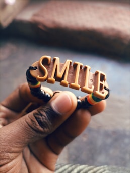 A hand holds a bracelet with the word 'SMILE' spelled out in raised, blocky letters. The bracelet features colorful elements, including beads in green, yellow, and red.
