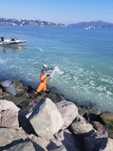 A person wearing orange overalls and a checked shirt is standing on rocky terrain next to the sea, casting a fishing net. A small motorboat floats nearby on calm waters with some distant hills and buildings visible on the shoreline.