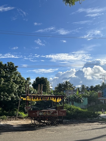 A small, wheeled food cart is positioned on the side of a road with a lush green background and trees. The cart is decorated with colorful images of fruits and has yellow triangular flags hanging from its canopy. In the background, several houses are visible amidst dense foliage, and the sky is clear with a few scattered clouds.