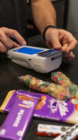 A person is using a payment terminal to complete a transaction, with hands holding a card and interacting with the device. Various snacks, including a bag of colorful gummy candies and chocolate bars in purple packaging, are placed on a dark countertop.
