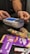 A person is using a payment terminal to complete a transaction, with hands holding a card and interacting with the device. Various snacks, including a bag of colorful gummy candies and chocolate bars in purple packaging, are placed on a dark countertop.