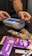 A person is using a payment terminal to complete a transaction, with hands holding a card and interacting with the device. Various snacks, including a bag of colorful gummy candies and chocolate bars in purple packaging, are placed on a dark countertop.
