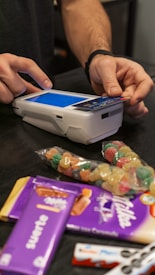 A person is using a payment terminal to complete a transaction, with hands holding a card and interacting with the device. Various snacks, including a bag of colorful gummy candies and chocolate bars in purple packaging, are placed on a dark countertop.