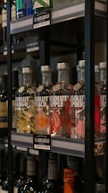 Several bottles of flavored vodka are displayed on a shelf in a store. The bottles are labeled with flavors such as mango, peach, apple, and raspberry. Each bottle has a unique colorful design representing its flavor. The price tags indicate the brand and cost in local currency.