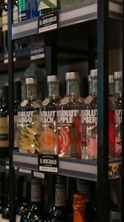 Several bottles of flavored vodka are displayed on a shelf in a store. The bottles are labeled with flavors such as mango, peach, apple, and raspberry. Each bottle has a unique colorful design representing its flavor. The price tags indicate the brand and cost in local currency.